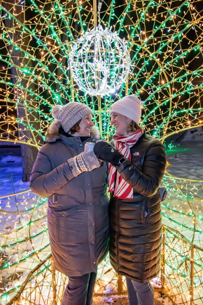 Two women toasting their beverages under illuminated holiday lights