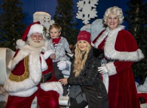 A little boy sits on Santa's lap, surrounded by his mother and Mrs. Claus